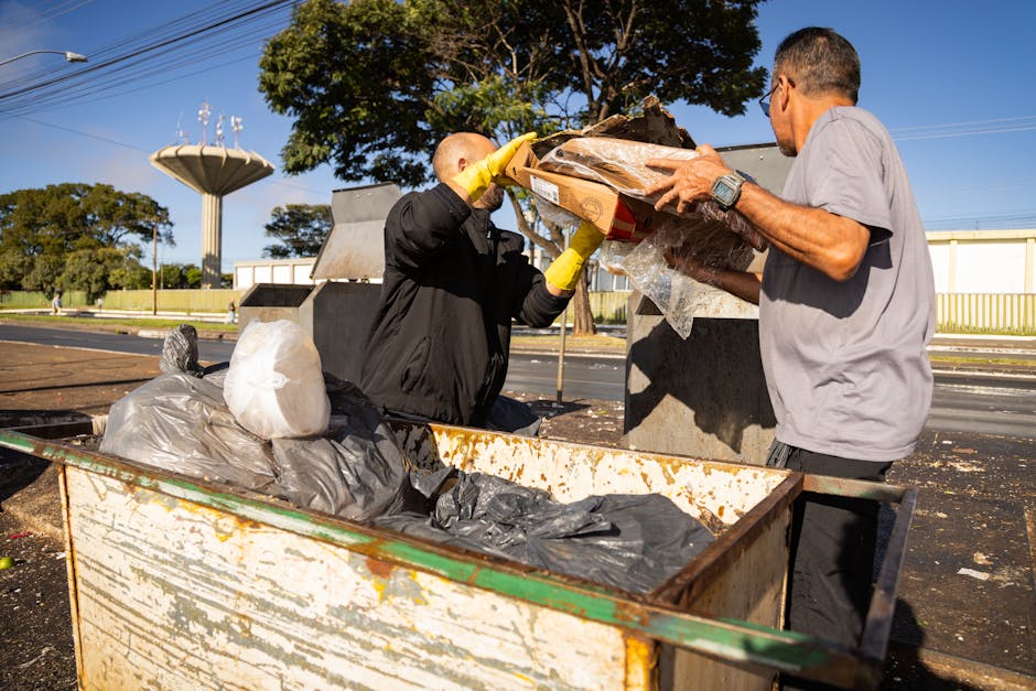 Two men are engaged in rubbish removal outdoors, standing next to a large open metal dumpster filled with black and grey plastic refuse bags, cardboard boxes, and loose debris. The man on the left, ba