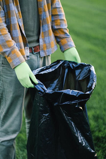 Two men are engaged in rubbish removal outdoors, standing next to a large open metal dumpster filled with black and grey plastic refuse bags, cardboard boxes, and loose debris. The man on the left, ba